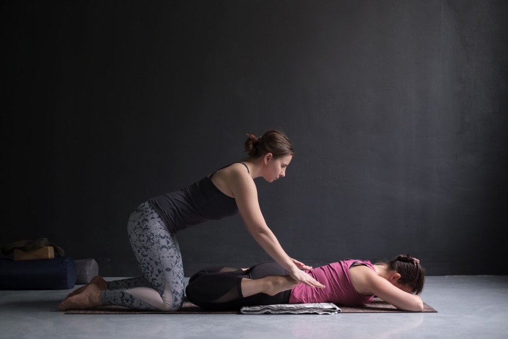 Coach de yoga qui aide une femme à faire la position de la grenouille aussi appelée bhekasana en Yoga.